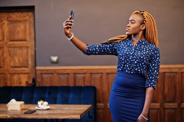 Portrait of beautiful young african business woman with dreadlocks, wear on blue blouse and skirt, posed in cafe and making selfie on phone.