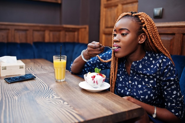 Ritratto di giovane e bella donna d'affari africana con i dreadlocks indossa una camicetta blu e gonna seduta in un bar con gelato e succo di ananas