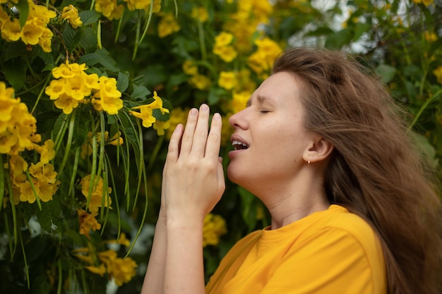 Photo portrait of beautiful young allergic woman is suffering from pollen allergy or cold on natural flower flowering tree background at spring or sunny summer day sneezes blowing her runny nose rubs eyes