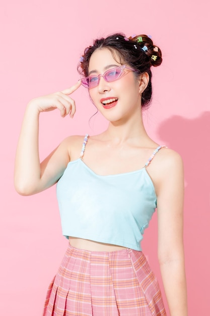 Portrait of beautiful young Asian happy woman in blue cute fashion dress wear sunglass smiling and posing on isolated pastel pink color background empty space