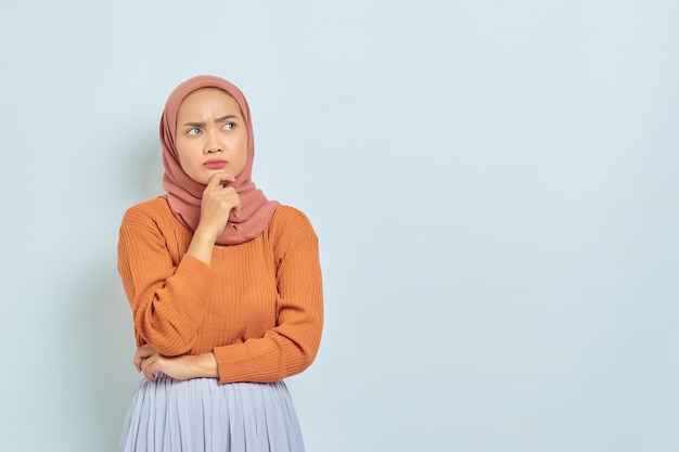 Portrait of beautiful young asian muslim woman in brown sweater
holding chin and looking seriously thinking question and looking at
copy space isolated on white background muslim lifestyle
concept