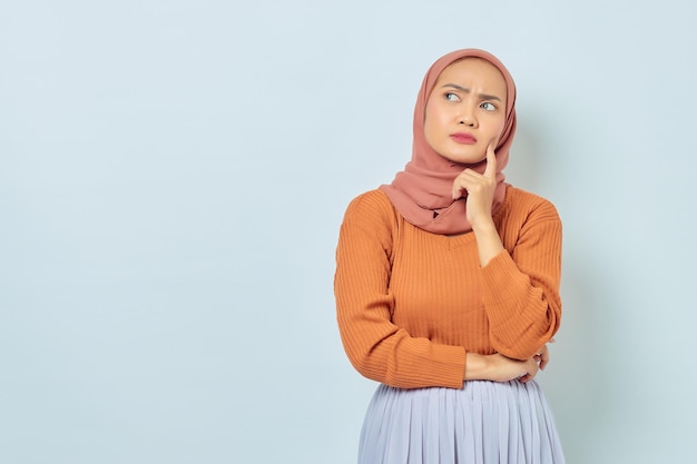 Portrait of beautiful young asian muslim woman in brown sweater\
looking serious thinking about question and looking at copy space\
isolated on white background muslim lifestyle concept