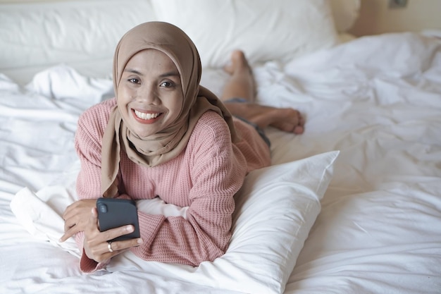 Portrait of a beautiful young Asian Muslim woman wearing hijab on bed holding mobile phone