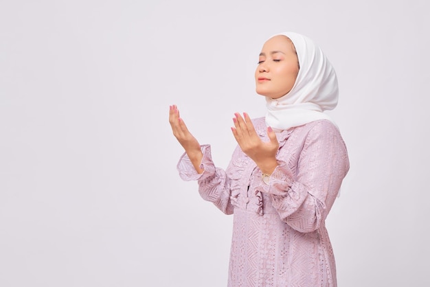 Portrait of beautiful young Asian Muslim woman wearing hijab and purple dress raising hands and praying to god isolated on white studio background