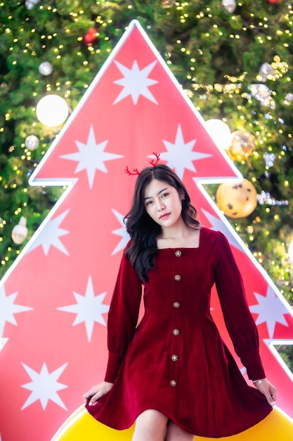 肖像画の美しい若いアジアの女性の赤いドレスの衣装とクリスマスツリーのクリスマスの挨拶の絵の小包の装飾明るい円形のボケの背景クリスマスと新年の装飾。