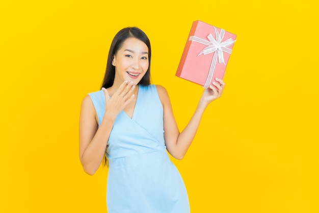 Sorriso di bella giovane donna asiatica del ritratto con confezione regalo rossa sulla parete di colore giallo