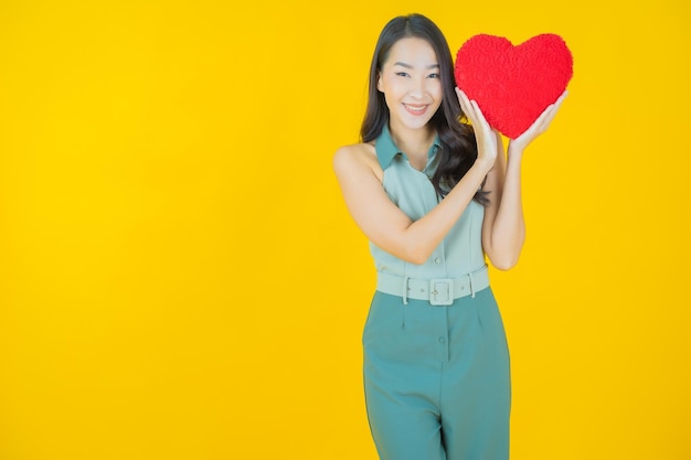 노란 벽에 심장 베개 모양으로 웃는 아름다운 젊은 아시아 여성의 초상화