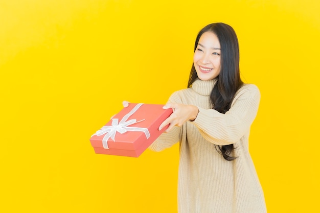 La bella giovane donna asiatica del ritratto sorride con il contenitore di regalo rosso sulla parete gialla