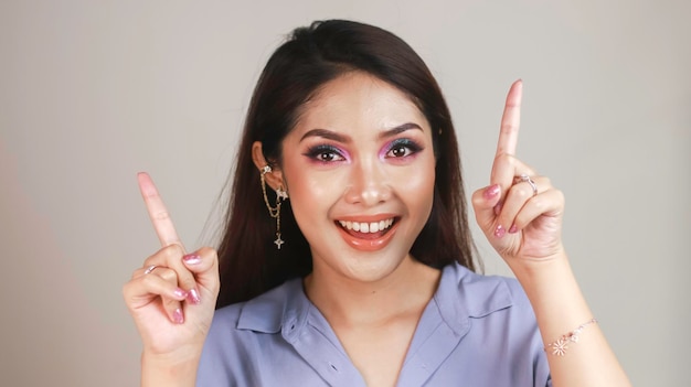 Portrait of beautiful young Asian woman smiling and pointing fingers upward at copy space isolated over white background