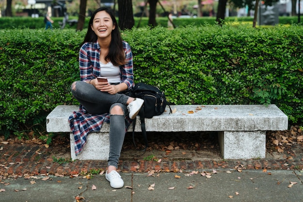 携帯電話を持ってベンチに座って笑顔の美しい若いアジアの女性の肖像画。陽気な女子大生は、オンラインで面白いチャットをしている携帯電話で笑っているカメラに顔を組んで足を組んだ。