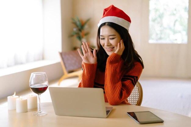 Портрет красивой молодой азиатки, улыбающейся, устраивая вечеринку по видеозвонку с друзьями и семьей, говоря привет, машет рукой на празднование Рождества в гостиной дома Зимние праздники Празднования онлайн