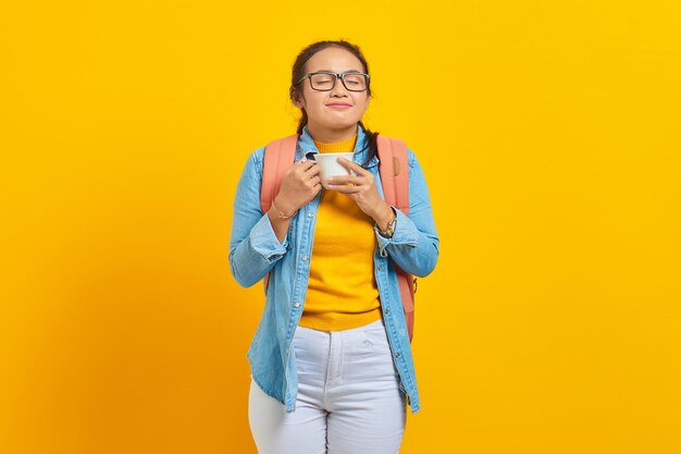 Photo portrait of beautiful young asian woman student in denim clothes with backpack while enjoying cup of coffee isolated on yellow background