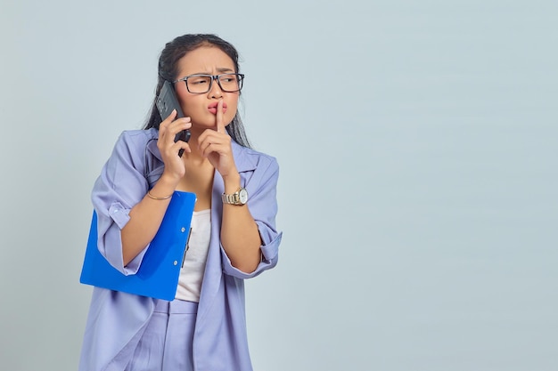 Portrait of beautiful young Asian woman talking on mobile phone and making silent gesture isolated on purple background