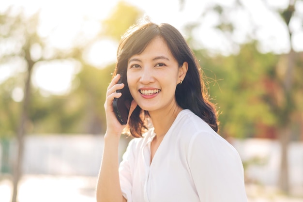 晴れた夏の日に屋外の自然の景色の周りにスマートな携帯電話を持つ美しい若いアジア女性の肖像画