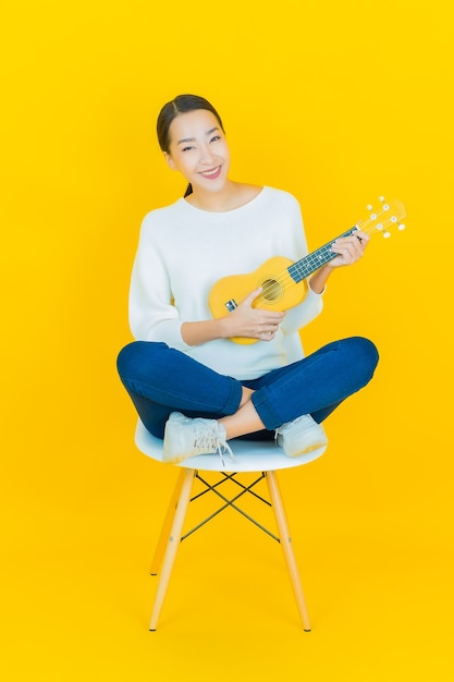 Женщина портрета красивая молодая азиатская с гавайской гитарой на желтом