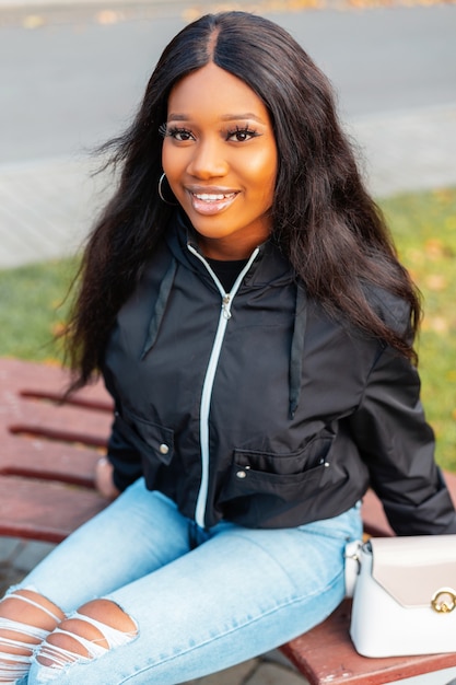 ブルージーンズとバッグとファッショナブルなカジュアルジャケットの笑顔で美しい若い黒人女性の肖像画は公園のベンチに座っています