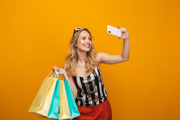 Portrait of a beautiful young blonde woman standing isolated over yellow background, taking a selfie