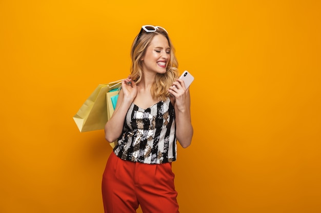 Портрет красивой молодой блондинки, стоящей изолированно на желтом фоне, с помощью мобильного телефона