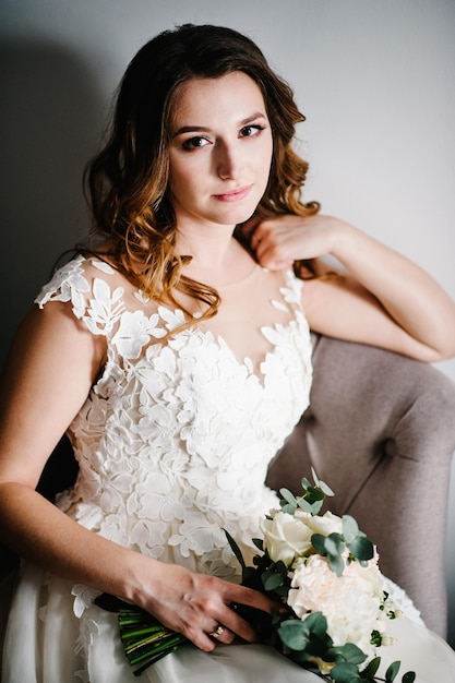 Portrait of a beautiful young bride in dress with wedding bouquet of flower on retro armchair. Tender happy emotion on the face.