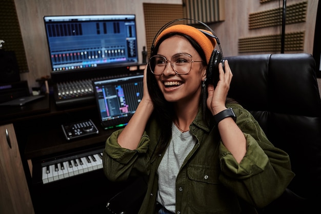 Photo portrait of beautiful young brunette female artist smiling aside adjusting headphones while sitting