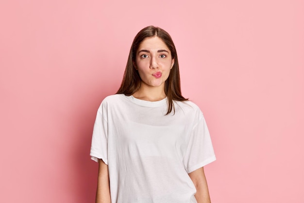 Portrait of beautiful young brunette girl posing in white tshirt with grimacing face against pink studio background Thoughtful look Concept of youth emotions facial expression lifestyle