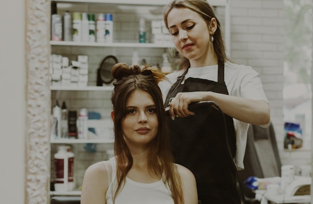 Portrait of a beautiful young brunette girl sitting in a chair in front of a mirror which the hairdresser twists her hair on a curling iron doing a wedding hairstyle closeup side view