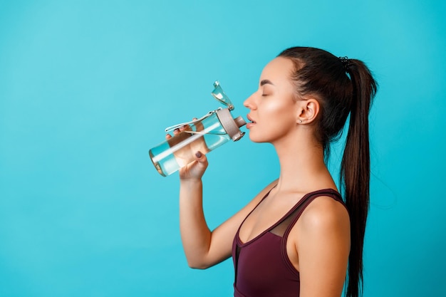 Portrait of a beautiful young brunette woman in a sportswear drinks water from a bottle on a blue background. space for text