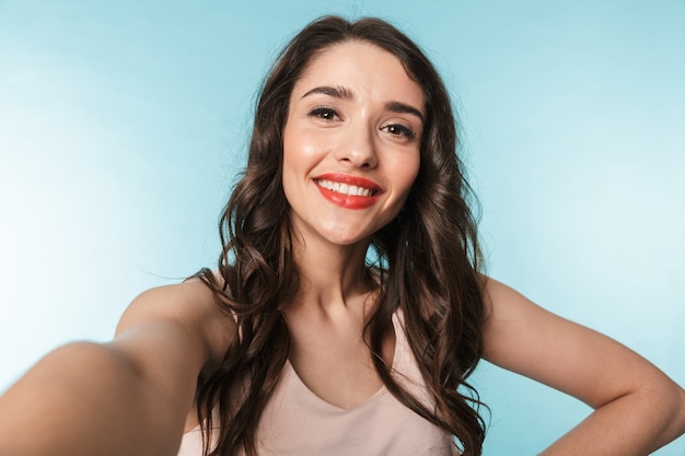 Portrait of a beautiful young brunette woman standing over blue, taking a selfie