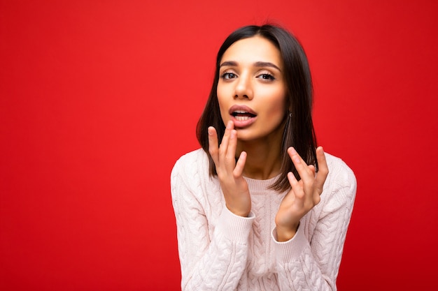 Portrait of beautiful young brunette woman wearing knitted jersey isolated over red background with free space and touching lips. Beauty concept