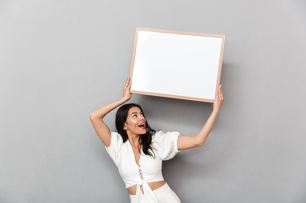 Portrait of a beautiful young brunette woman wearing summer outfit standing isolated over gray wall, showing blank board