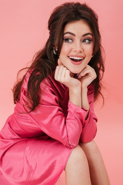 Portrait of a beautiful young brunette woman with bright makeup wearing fashionable clothes sitting isolated over pink wall