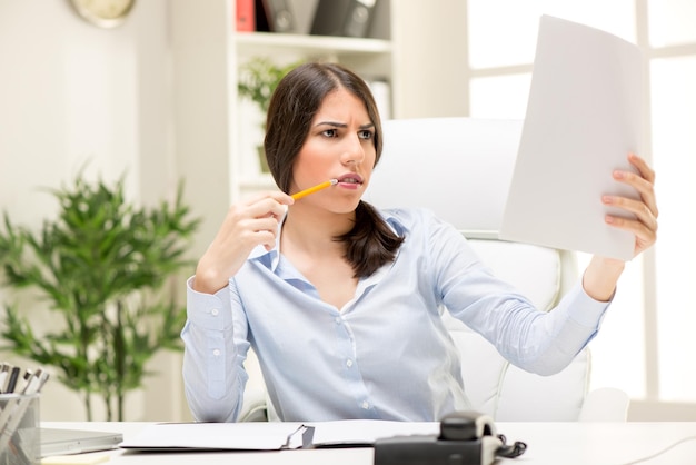 Portrait of a beautiful young businesswoman with problems. She is sitting in the office, looking at document, biting pen and thinking.