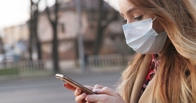 Portrait of beautiful young Caucasian woman in medical mask using her smartphone outdoor. Pretty girl texting message on phone during pandemic at street. Pandemic concept, coronavirus epidemic.