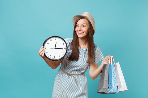 Ritratto bella giovane donna caucasica in abito estivo, cappello di paglia che tiene pacchetti borse con acquisti dopo lo shopping, orologio rotondo isolato su sfondo blu pastello. copia spazio per la pubblicità.