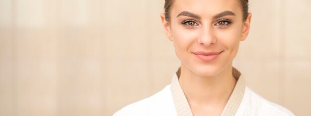 Ritratto di una giovane e bella donna caucasica con i capelli scuri che indossa accappatoio bianco che guarda l'obbiettivo e sorridente su uno sfondo beige