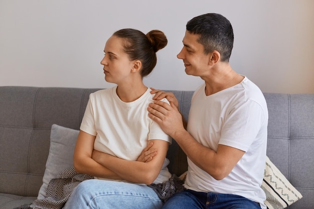 Portrait of beautiful young couple is having a quarrel while sitting on sofa at home, wife turning her body and being offended, husband with smile trying to talk and reconcile.