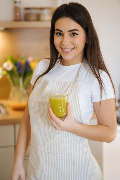 Portrait of beautiful and young female in apron standing in the kitchen woman hold glass of green