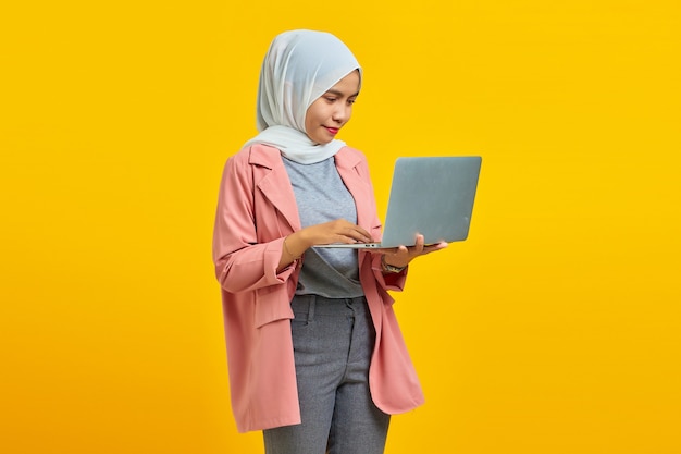 Portrait of beautiful young female college student with laptop smiling wearing pink jacket isolated on blue background