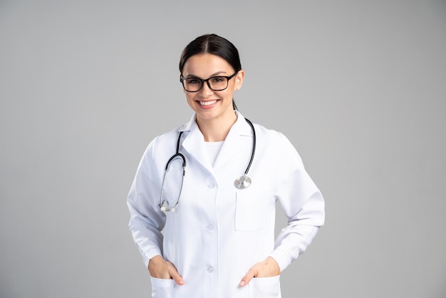 Portrait of beautiful young female doctor in white medical jacket isolated on grey background. Brunette woman cosmetologist smiling at the camera