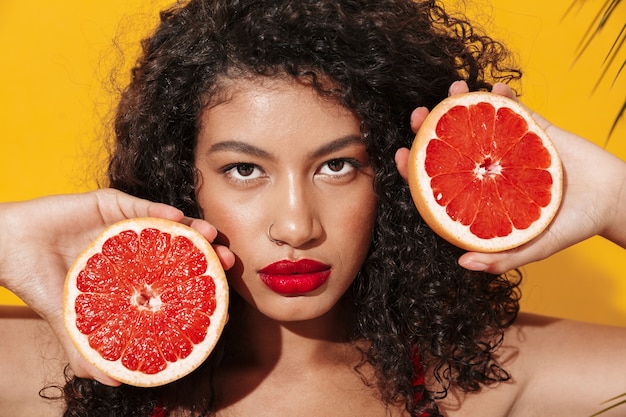 Portrait of a beautiful young fit woman wearing bikini standing isolated over yellow exotic wall, posing with halved grapefruit