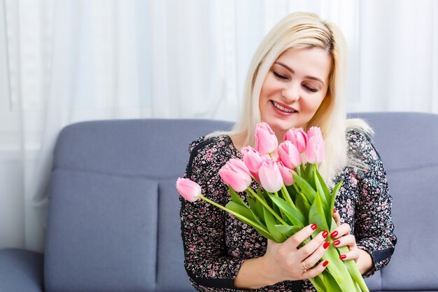 Portrait of a beautiful young girl in dress holding big bouquet of tulips at home on the couch