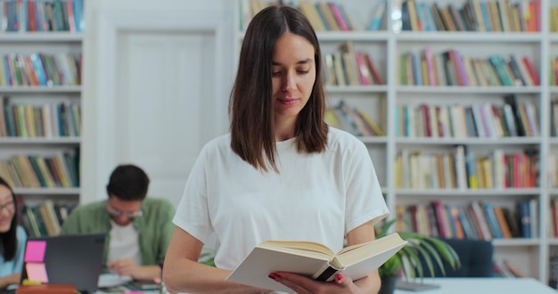 Ritratto di giovane e bella ragazza in possesso di un libro sullo sfondo dello scaffale della biblioteca guardando la fotocamera concetto di istruzione universitaria