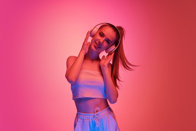 Portrait of beautiful young girl posing listening to music in\
headphones isolated over pink background in neon light