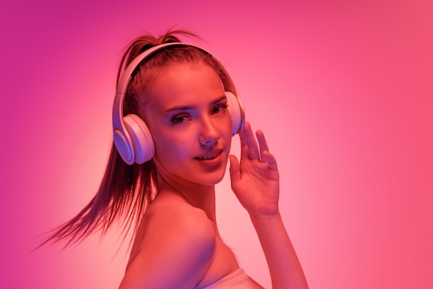 Portrait of beautiful young girl posing listening to music in headphones isolated over pink background in neon light