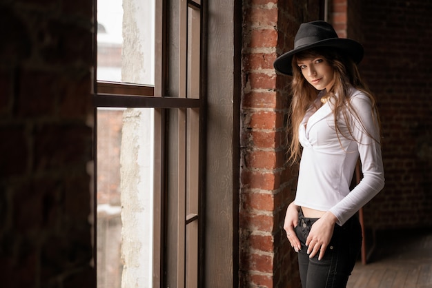 Portrait of beautiful young girl in white shirt and hat by the window