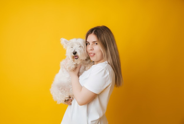 Portrait of a beautiful young girl with a dog in her arms on a yellow background the girl hugs her pet west highland white terrier copy space