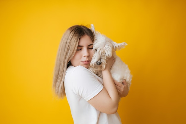 Portrait of a beautiful young girl with a dog in her arms on a yellow background the girl hugs her pet west highland white terrier copy space