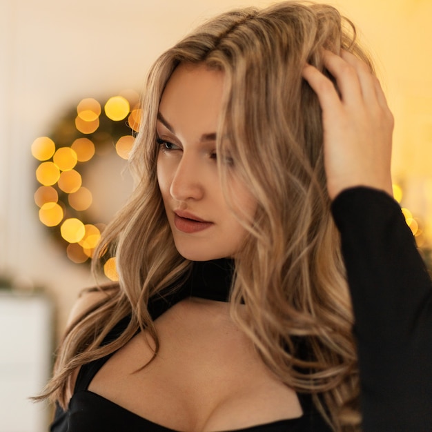 Portrait of a beautiful young girl with a hairstyle in a black elegant dress on a background of yellow bokeh lights. Christmas party and holidays