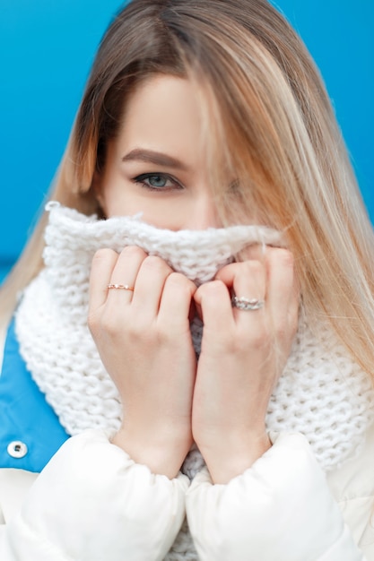 Portrait of a beautiful young happy girl with blue eyes with a knitted stylish scarf and a white winter jacket on blue outdoors