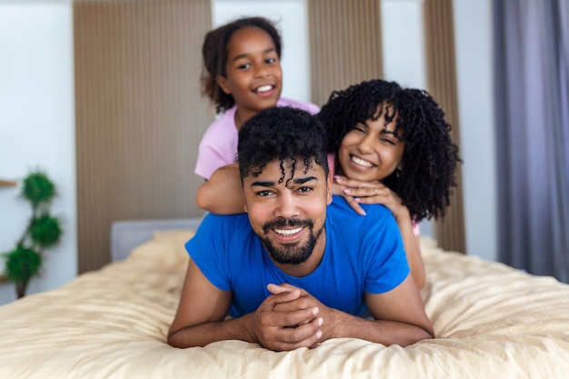 Портрет красивой молодой матери, отца и их дочери, смотрящих в камеру и улыбающихся, лежащих на постели и опирающихся друг на друга